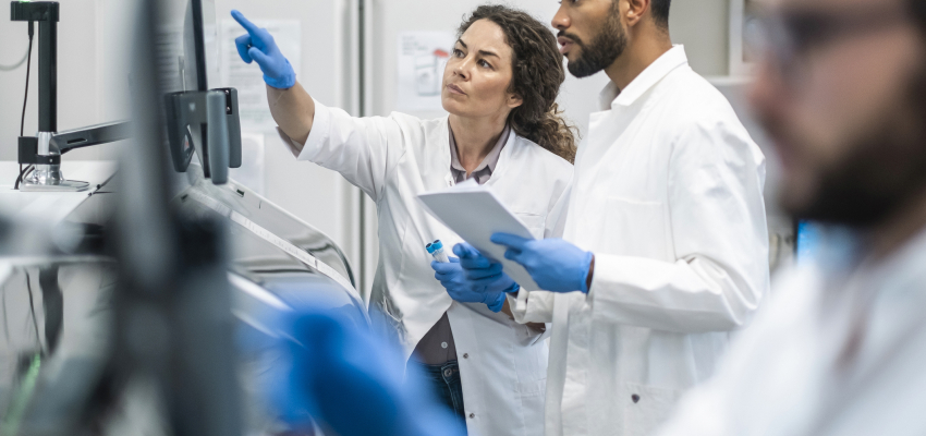 Two scientists in lab coats and gloves examining a computer screen in a laboratory.