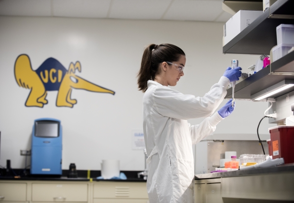 A student wearing a white coat doing research in a lab with Peter the anteater painted on the wall in the background