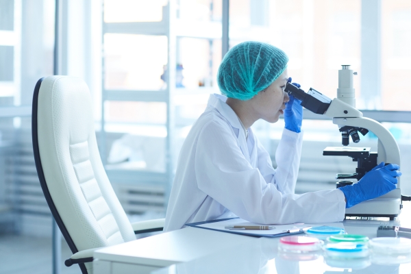 Researcher in protective gloves and cap looking in microscope while sitting at a lab desk.
