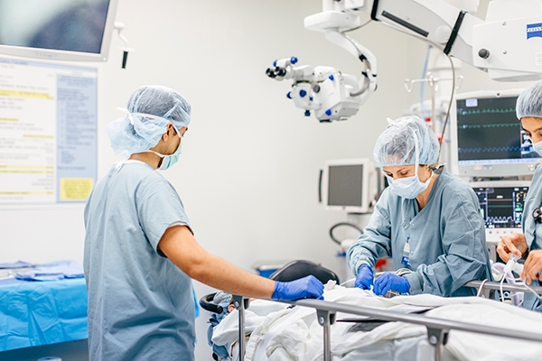 A trainee watches two doctors provide anesthesia to a patient on an operating table