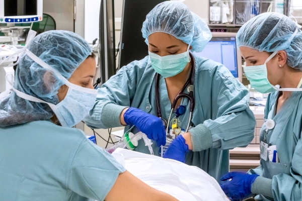 The image shows three healthcare workers in a medical setting, likely a hospital or operating room. They are wearing blue surgical scrubs, hair nets, and protective masks. They are gathered around a patient, whose head is not visible, as they prepare or perform a medical procedure. One of the workers in the center is holding medical instruments and wearing a stethoscope around the neck. All are wearing blue gloves, indicating they are maintaining a sterile environment. In the background, there are medical e