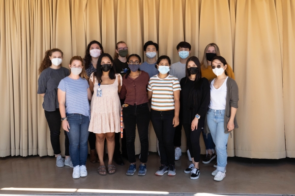 Group photo of 12 people wearing masks standing in front of a yellow stage curtain.