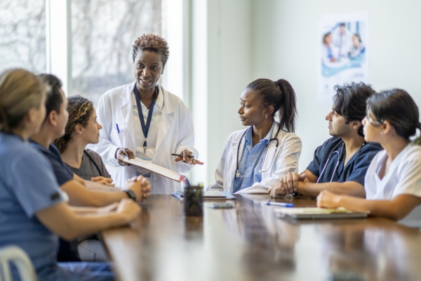 A small group of diverse nursing students sit around a boardroom table as they listen attentively to their teacher and lead doctor. They are each dressed in medical scrubs and sitting with papers out in front of them. The doctor is holding out a clipboard with a document on it as she reviews it with the group.