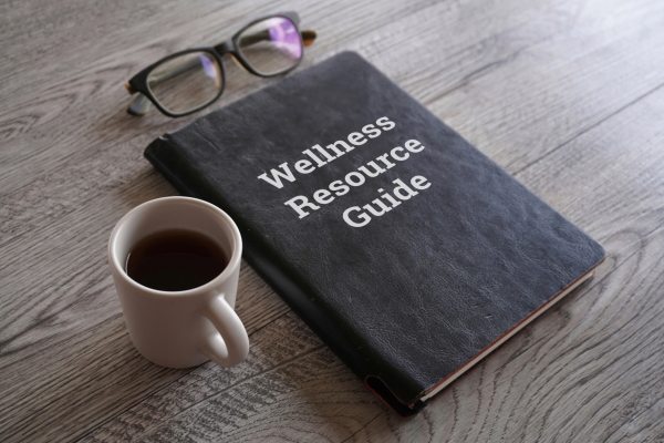 a photo of a black book that says Wellness Resource Guide on the cover. There is also eye glasses and a cup of coffee on a wooden table.