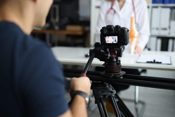 Videographer in foreground is filming a person in a white coat for online education.