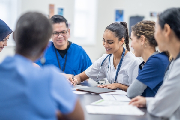 A small group of medical students gather around a table as they meet together to discuss patient cases. They are each dressed professionally and have files scattered between them for reference.