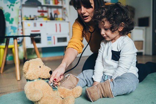 Pediatrician kneeling next to toddler pretending to listen to his teddy bear's heart