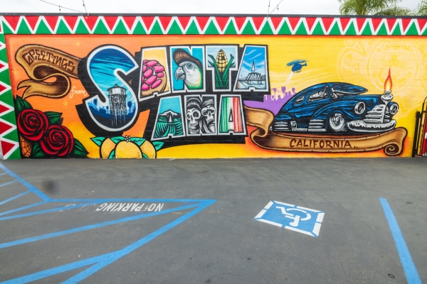 Colorful postcard-style mural that reads “Greetings from Santa Ana, California,” with imagery including a parrot, lowrider car, citrus, roses, and local landmarks.