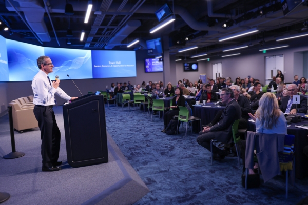 Dean Michael Stamos presents at the 2025 Faculty Research Retreat at a podium to an audience in a modern conference room with large screens in the background.