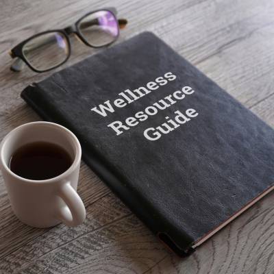 a photo of a black book that says Wellness Resource Guide on the cover. There is also eye glasses and a cup of coffee on a wooden table.