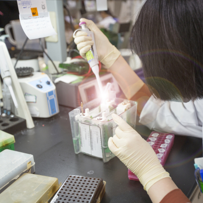 A scientist in a lab coat and gloves uses a micropipette to load a sample into a gel electrophoresis apparatus.