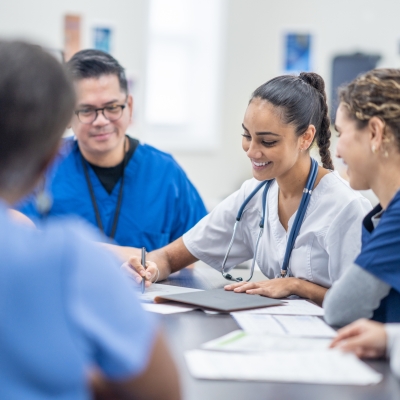 A small group of medical students gather around a table as they meet together to discuss patient cases. They are each dressed professionally and have files scattered between them for reference.