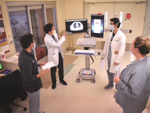 Two doctors in white coats pointing to medical imaging on screens, explaining to other healthcare workers