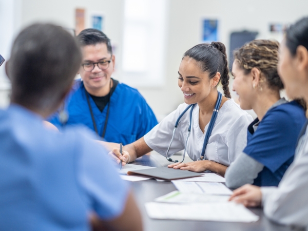 A small group of medical students gather around a table as they meet together to discuss patient cases. They are each dressed professionally and have files scattered between them for reference.