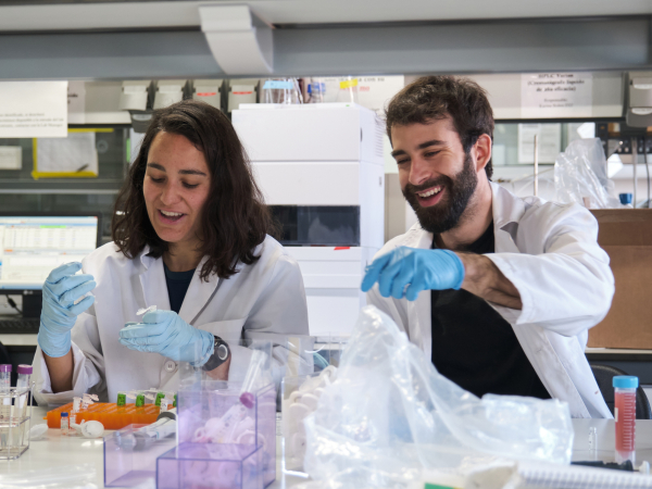 Two laboratory workers in white lab coats and blue gloves work with containers at a lab bench.
