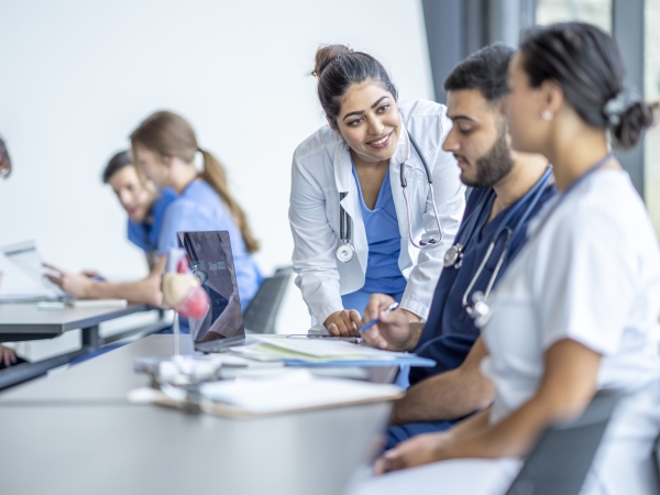 Professor in scrubs leans into desk where students wearing scrubs are seated.