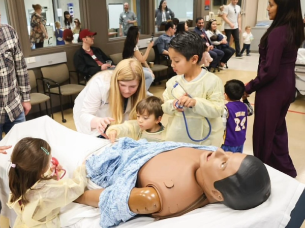 Several children gather with Alicia Wells around a patient care mannequin on a bed.