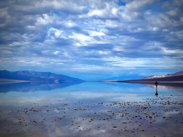 A peaceful lake mirroring billowy clouds in the sky. Mountains are in the distance, and a lone person stands at the edge of the lake.
