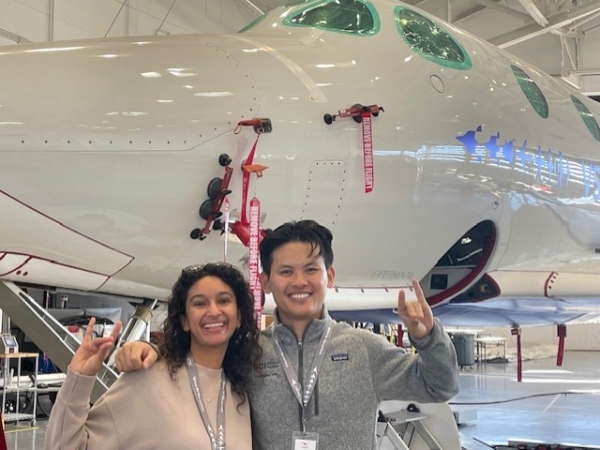 Kaveri Curlin and Christopher Yang stand in front of a plane, doing the UCI "zot" sign with their hands.