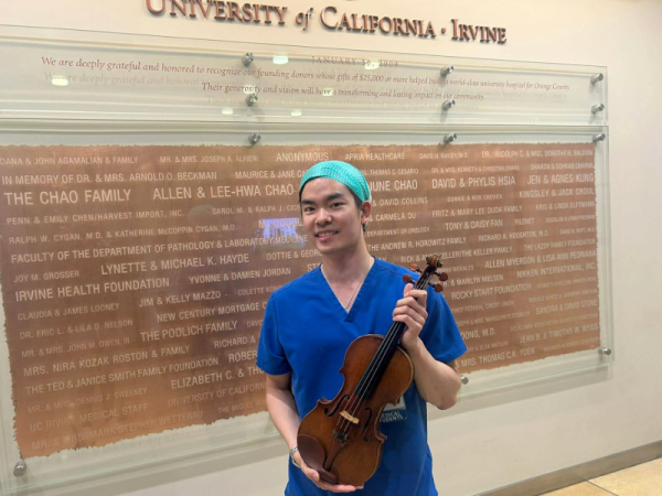Frank Lee wearing scrubs and holding a violin as he stands in front of a University of California, Irvine sign inside a hospital