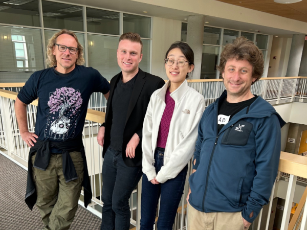 The four researchers stand together, leaning against a railing inside a building.
