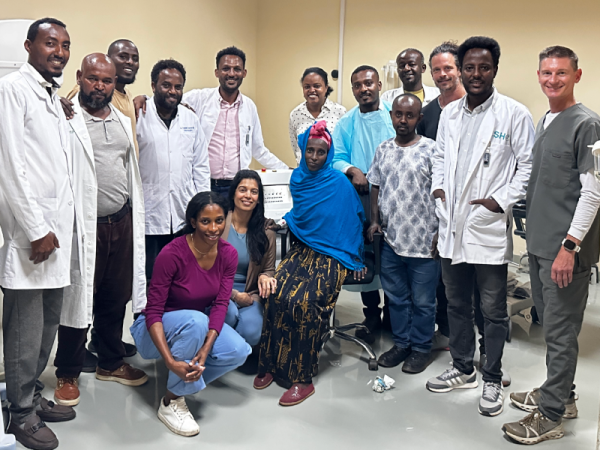 A patient sits in a chair, surrounded by a group of 14 people, many in white coats and medical scrubs