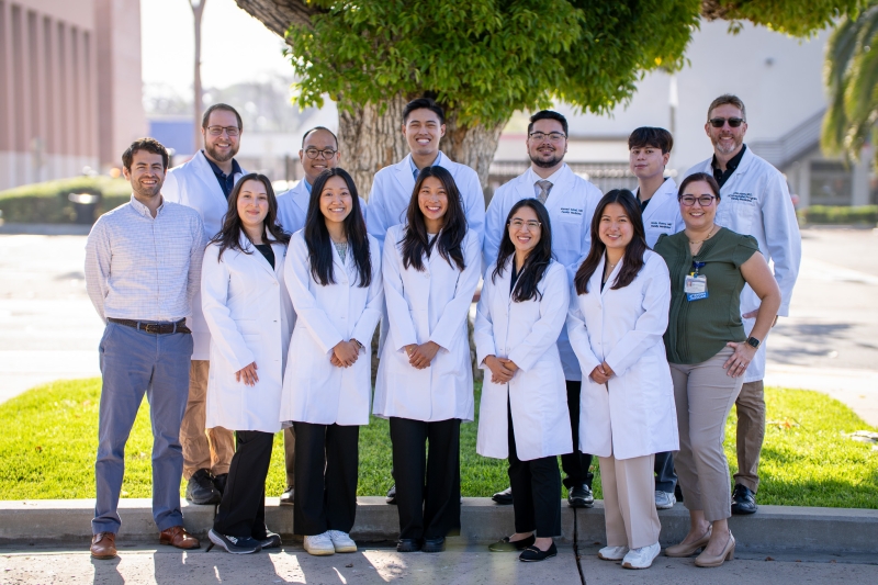 A group of twelve people, mostly in white lab coats, smiling outdoors in front of a tree.