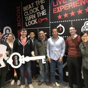 Eight people at an escape room event. One person holds a cutout of a detective while another holds a cutout of a key that says Exodus Escape Room. Behind them, on the wall, it says: Beat the clock & turn the lock.