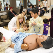 Several children gather with Alicia Wells around a patient care mannequin on a bed.