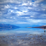 A peaceful lake mirroring billowy clouds in the sky. Mountains are in the distance, and a lone person stands at the edge of the lake.