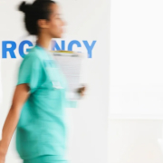 Motion-blurred healthcare worker in turquoise scrubs walking past an “EMERGENCY” sign in a bright hospital corridor.