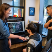 Two people stand next to someone sitting in front of a computer screen. The screen shows an atomic-resolution X-ray crystal structure of a protein.