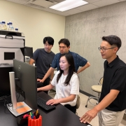 Sunhee Jung sits at a computer as the others stand around her, looking at the monitor.