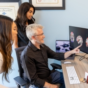 Three people looking at two different monitors. One shows three graphics of a brain, and the other shows the outline of a person’s head, neck and back, with various points all over the body.