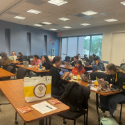 Students in a classroom with laptops and study materials, engaging in activities at wooden desks.