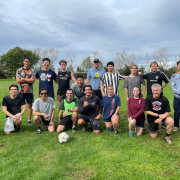 A group of 17 people, in athletic attire, pose on a grassy field with a soccer ball.
