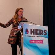 Sophie Zaaijer speaking at a podium with a sign that says, “Health Equity & Research Summit (HERS).”