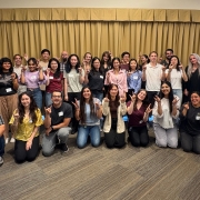 A group of people posing indoors with beige curtains in the background, many holding the "UC Irvine Anteater Zot Zot" hand sign.