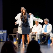 Medical student puts on white coat while on stage with faculty and staff. 