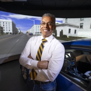 Federico E. Vaca stands next to a car door, in front of a large wrap-around screen showing a city road. 