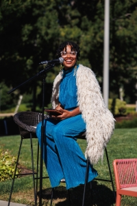 reelaviolette sits on a stool in front of a microphone, smiling and holding a book of poetry.