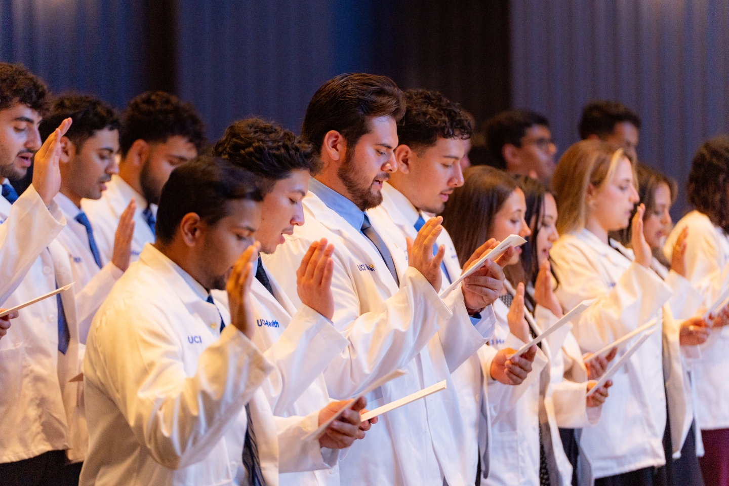 Students stand wearing their white coats, right hand raised, reciting the Hippocratic Oath.