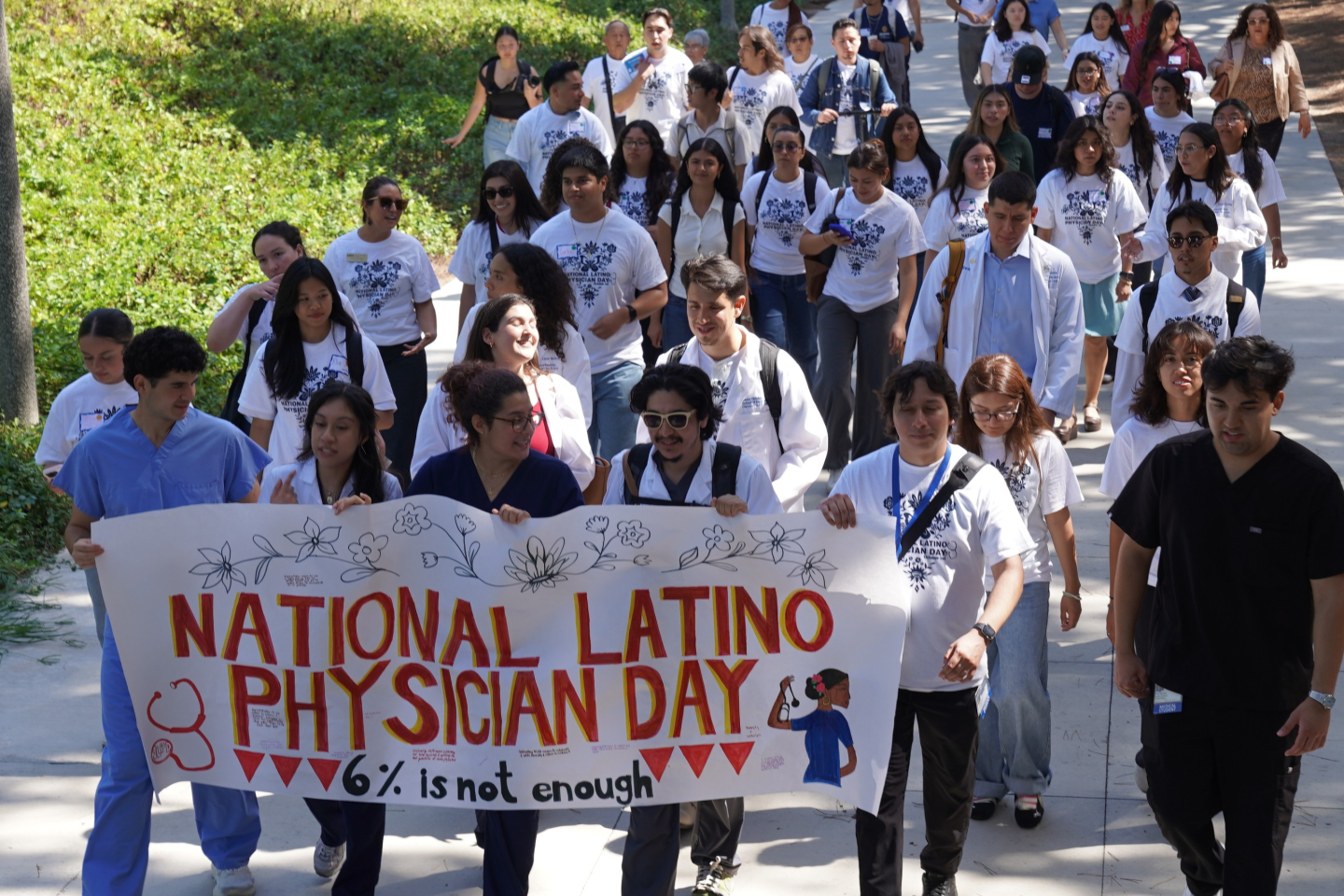 A large group of students, most in matching t-shirts, walk along a pathway. At the front, five students hold a banner that reads: "National Latino Physician Day: 6% is not enough." 