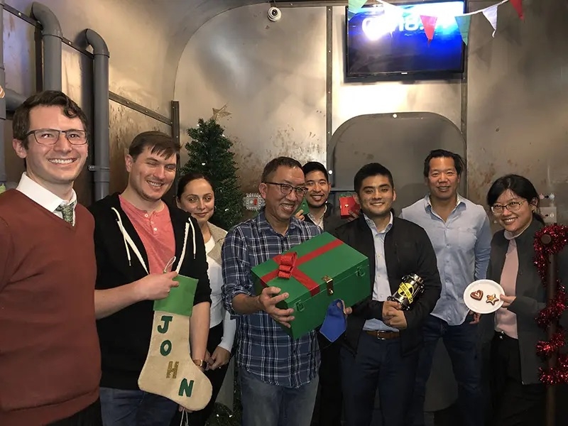 Eight people inside an escape room. One person holds a stocking that says "John." Another holds a green box with a red ribbon and a lock. One person holds a silver contraption. One person has a plate of two cookies.