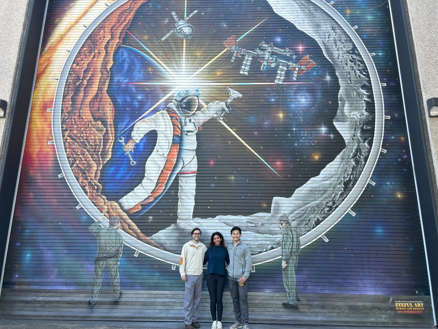 Sahin, Curlin, and Yang stand in front of a huge mural of an astronaut in space.