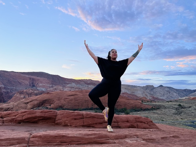 Glover stands on a rock, arms outstretched for her yoga pose, with mountains and sky in the backdrop