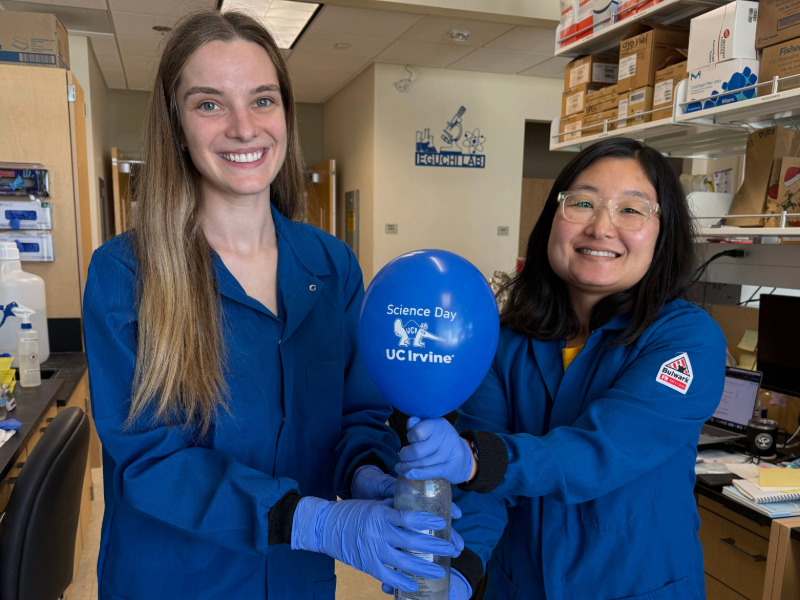 Asuka and Michelle stand in lab coats, with gloves on, holding a plastic water bottle with a blown-up balloon at the top that says Science Day UC Irvine.