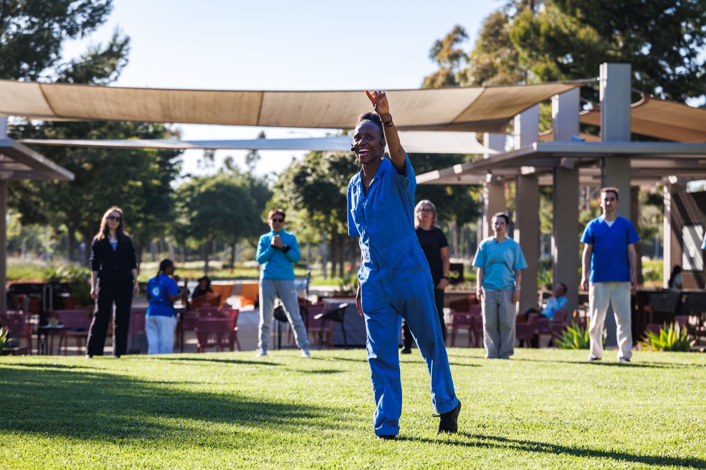 Ama Wray stands in the middle of a grass area, arm outstretched. Participants are in a circle around her.
