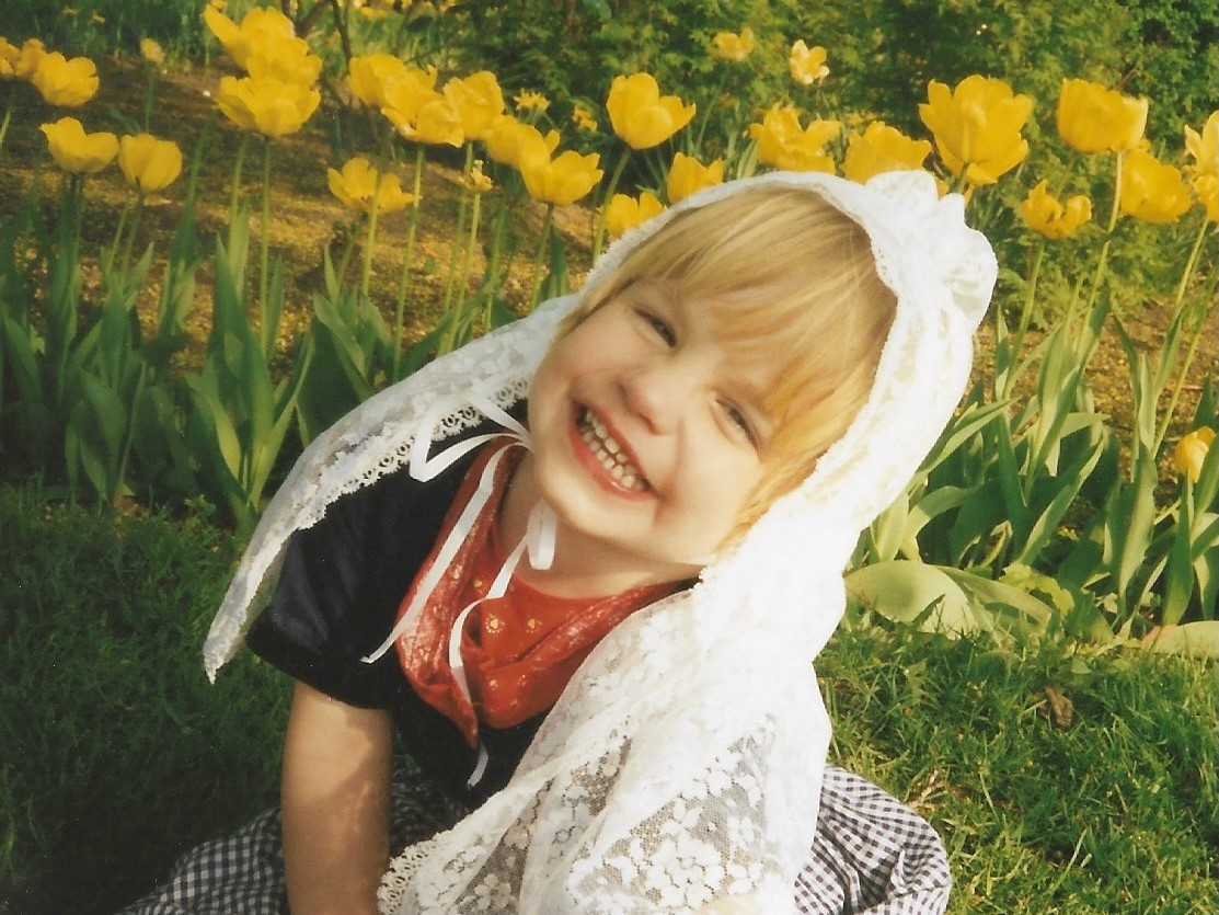 Cassandra Kooiker as a toddler, sitting in grass with flowers behind her.