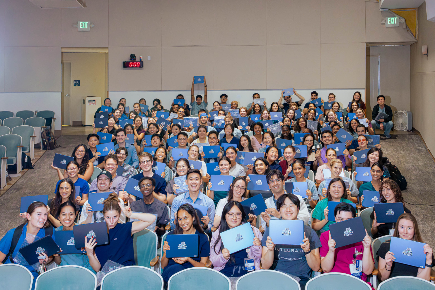 Medical students hold up their MacBook Neos, branded as ZOTbooks, in a lecture hall.